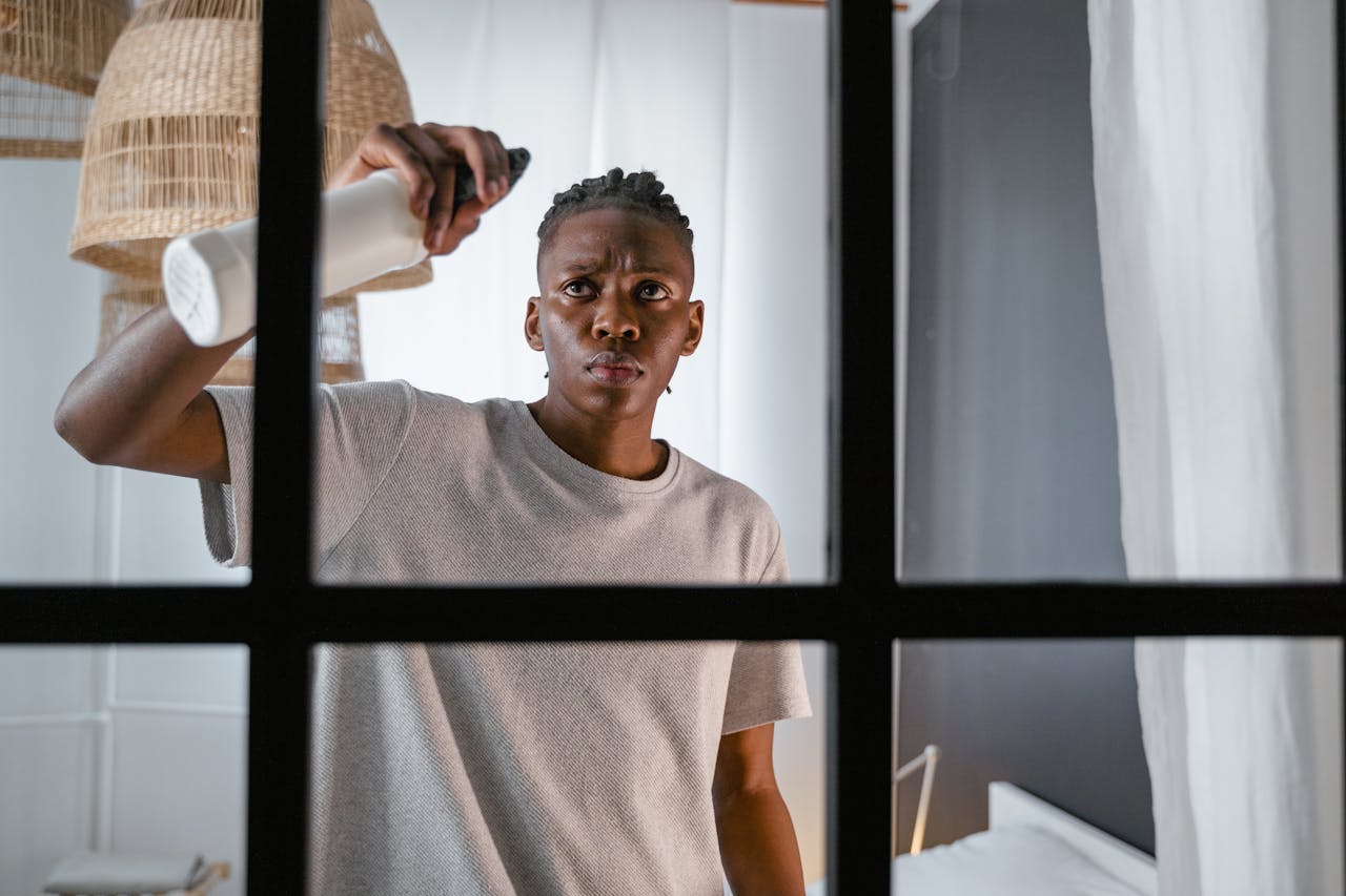 A young man cleaning a window indoors with a spray bottle, focusing on household chores.